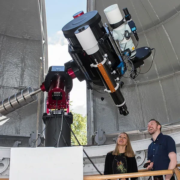 Two people looking up at a large telescope under a partially open domed roof..