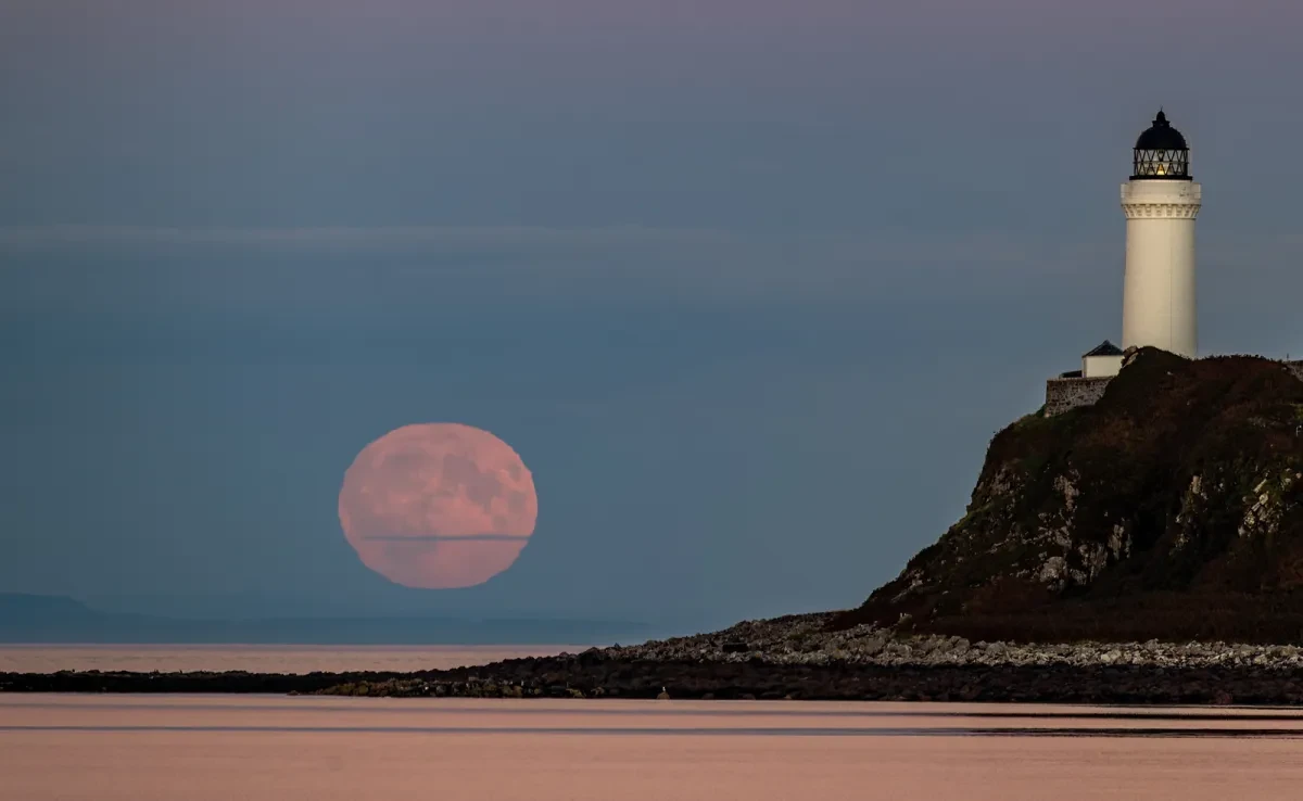 Photo showing beach landscape with rocky rise with lighthouse on top on the right hand side, and a low, large orange Moon on the left side