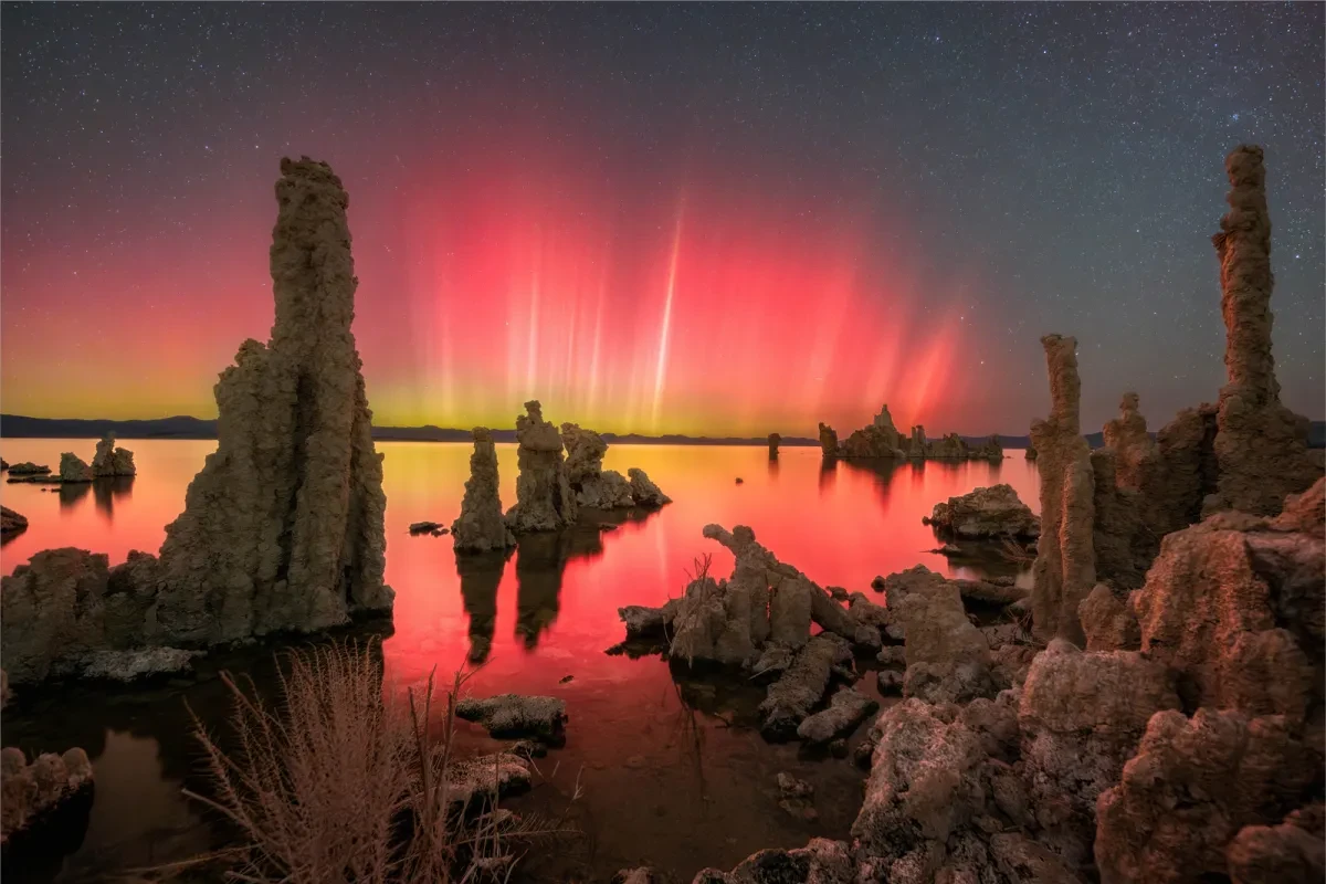 A dramatic red aurora display appears over a lake, with a rock formation in shadow in the foreground