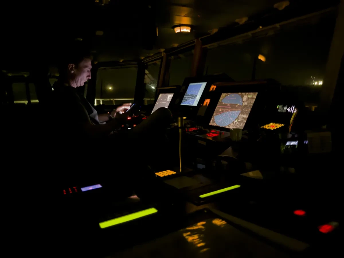 Night time view of the bridge of a container ship. The space is dark save for some dimly lit navigation screens and controls. A man is sat looking at a screen