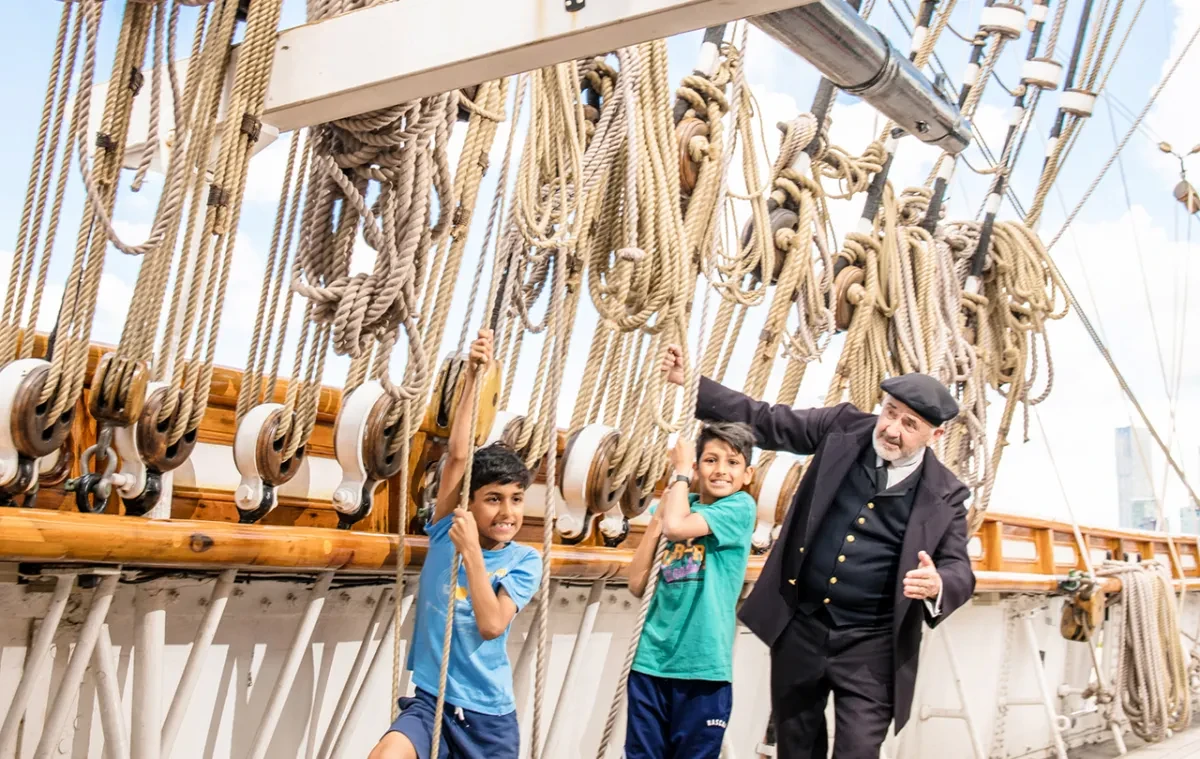Two children pull on Cutty Sark's ropes, alongside a man in a captain costume.