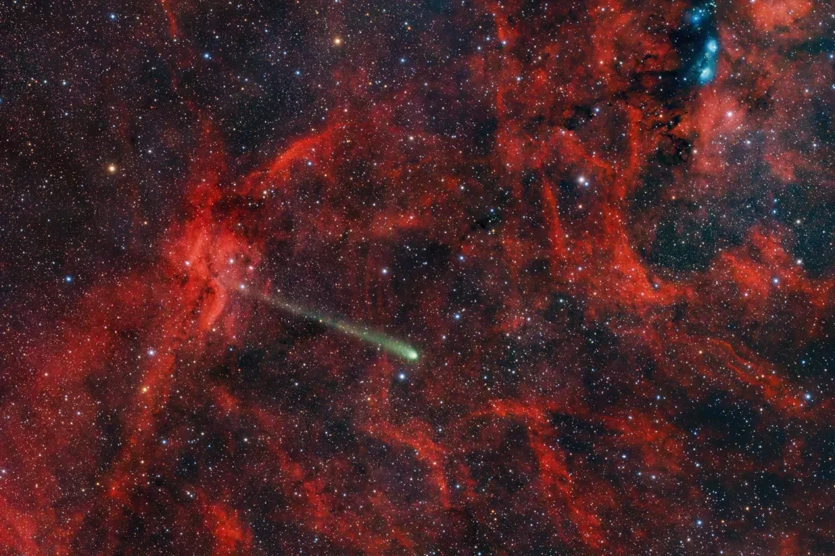 A green comet streaks in front of a red nebula.