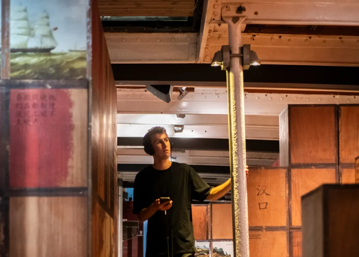 A young man walks amongst stacks of crates in the hold of a ship. He is wearing headphones and holding his phone, and appears to be listening intently