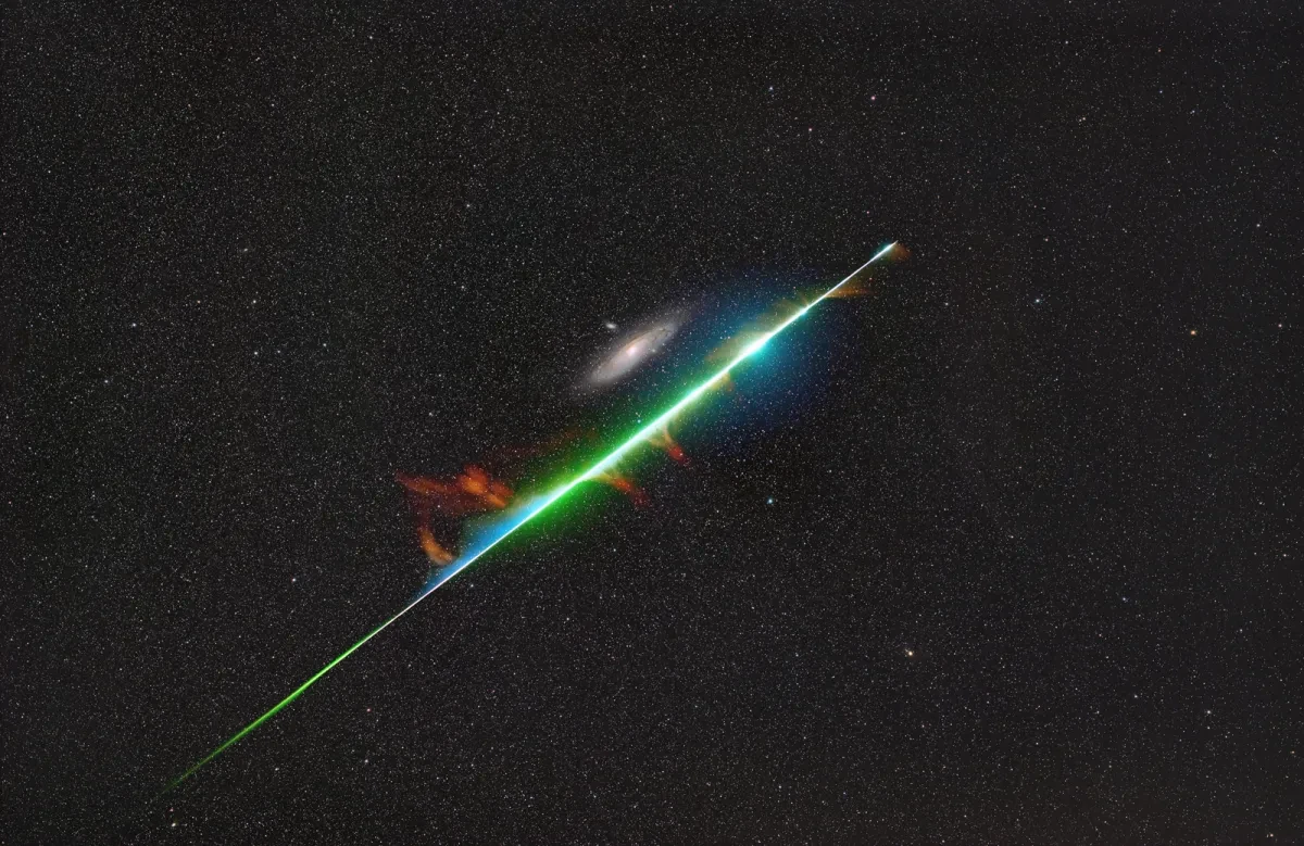 The very bright blue and green line of a fireball cuts through the starry sky next to a galaxy.