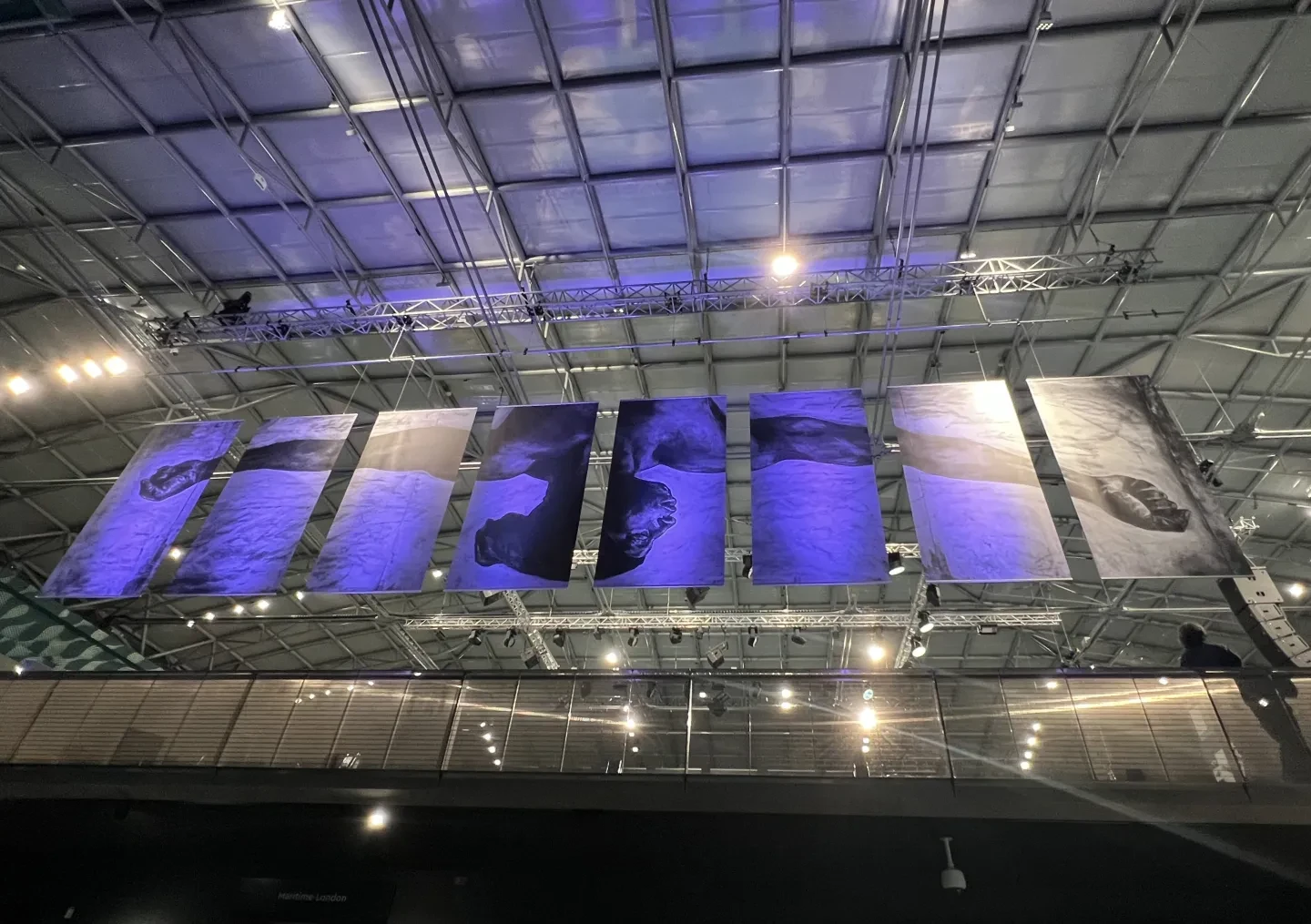 8 hanging banners with a illustration made by pencil of the upper body of an Afro-Caribbean male with arms spread wide upside down. The banners are lit up by blue lights in the National Maritime Museums' Ocean Court