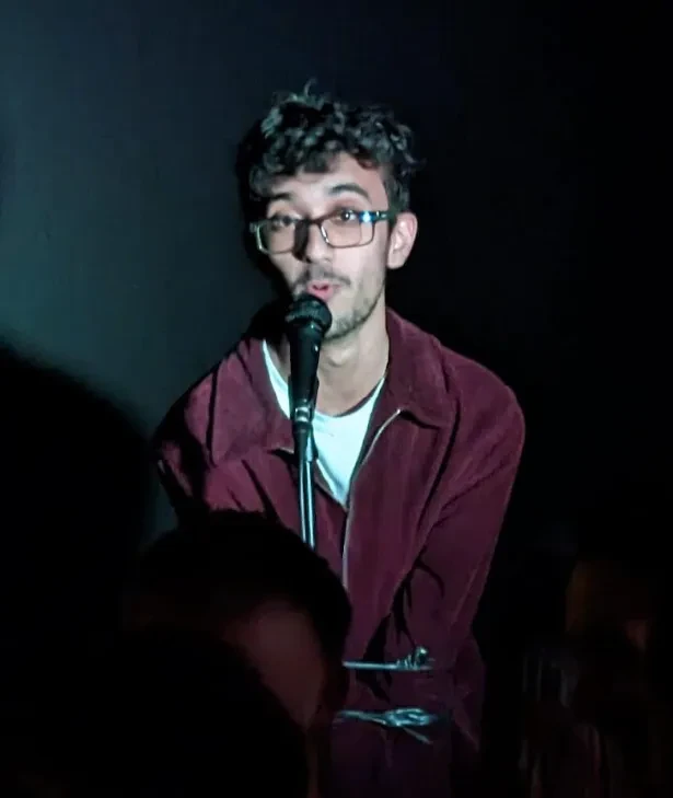 Aman, dark curly hair, wearing glasses face onto the camera. Speaking into the microphone, wearing a white top and red jacket 