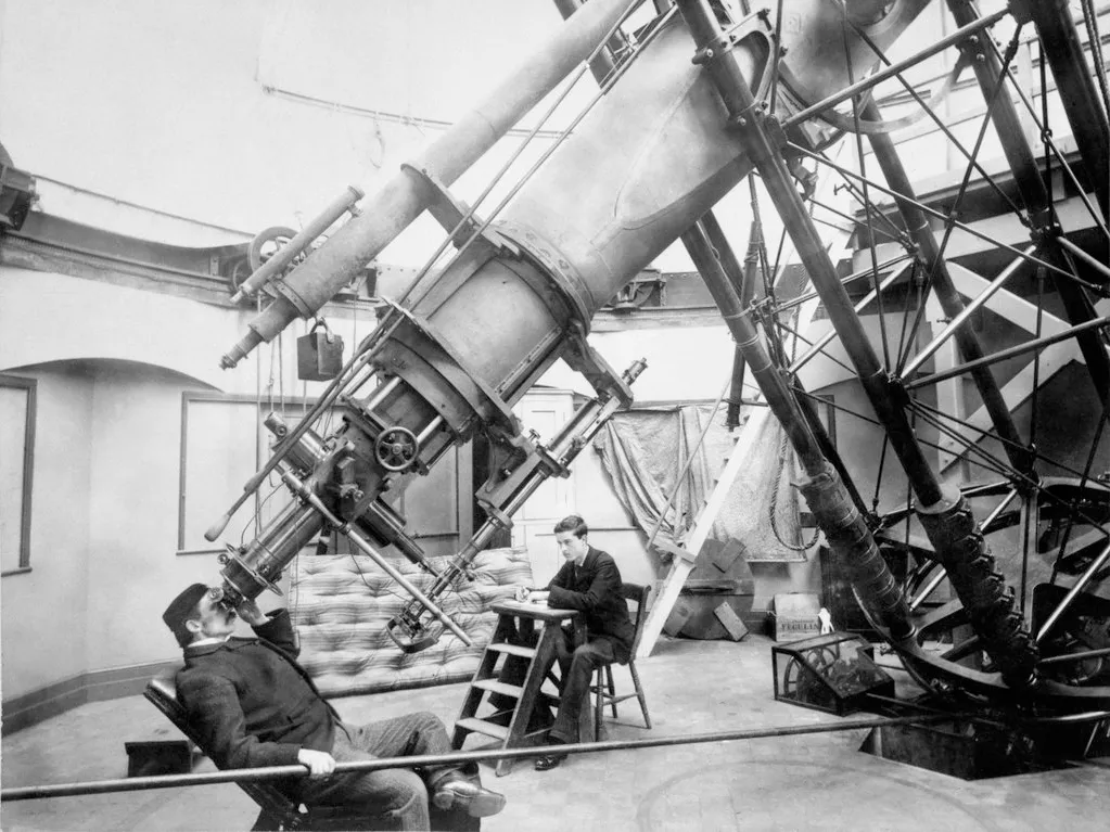Black and white photograph of an astronomer using the Great Equatorial Telescope at Royal Observatory Greenwich. He is leaning back on a small chair, his eye to the eyepiece of a giant telescope. Another man is sat at a bench behind