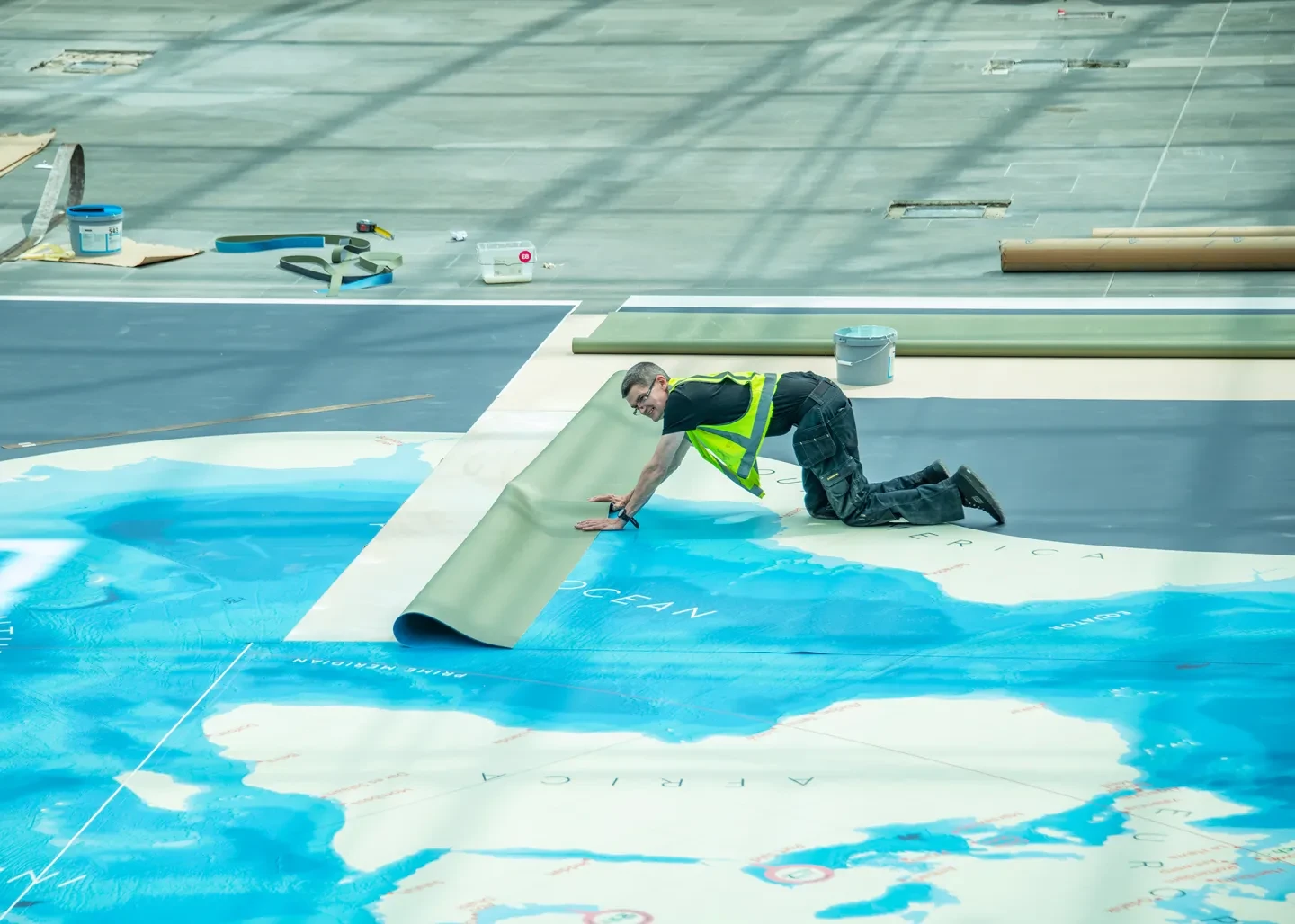 A person in a yellow hi-vis jacket rolls out a section of a large floor map of the world
