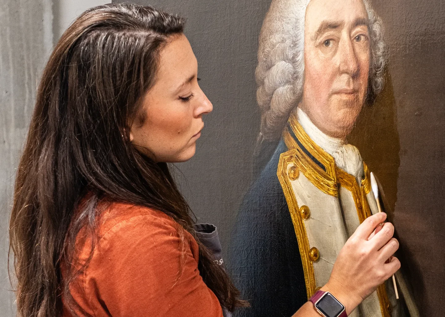A female paintings conservator works delicately on an oil painting of a man in historic naval uniform