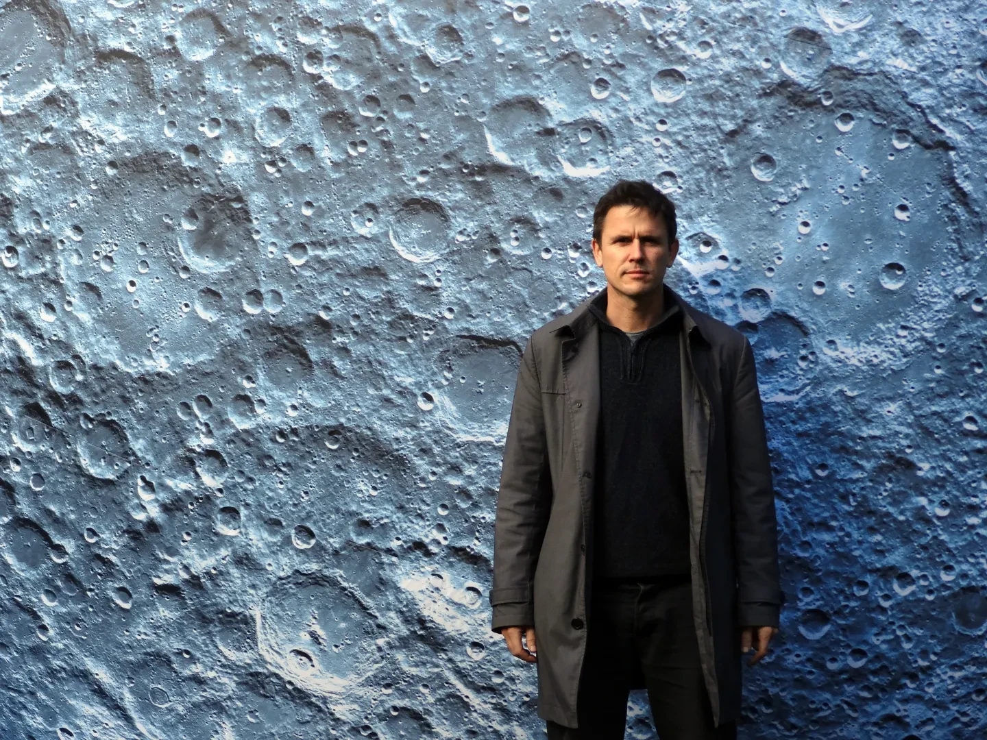 Luke Jerram stands in front of his earlier work, Museum of the Moon, wearing a brown coat, a dark top and a determined expression on his face