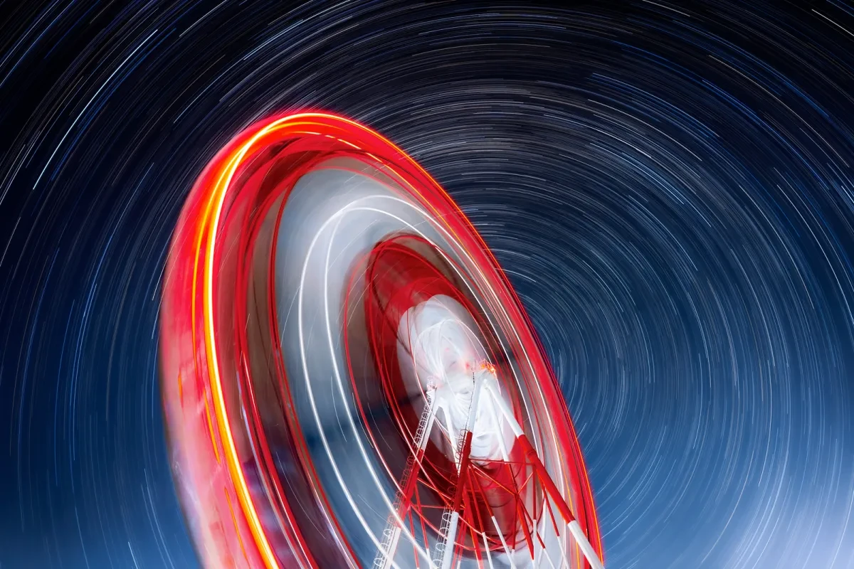 Image showing long exposure from ground level looking up at a red and white ferris wheel with circular star trails behind