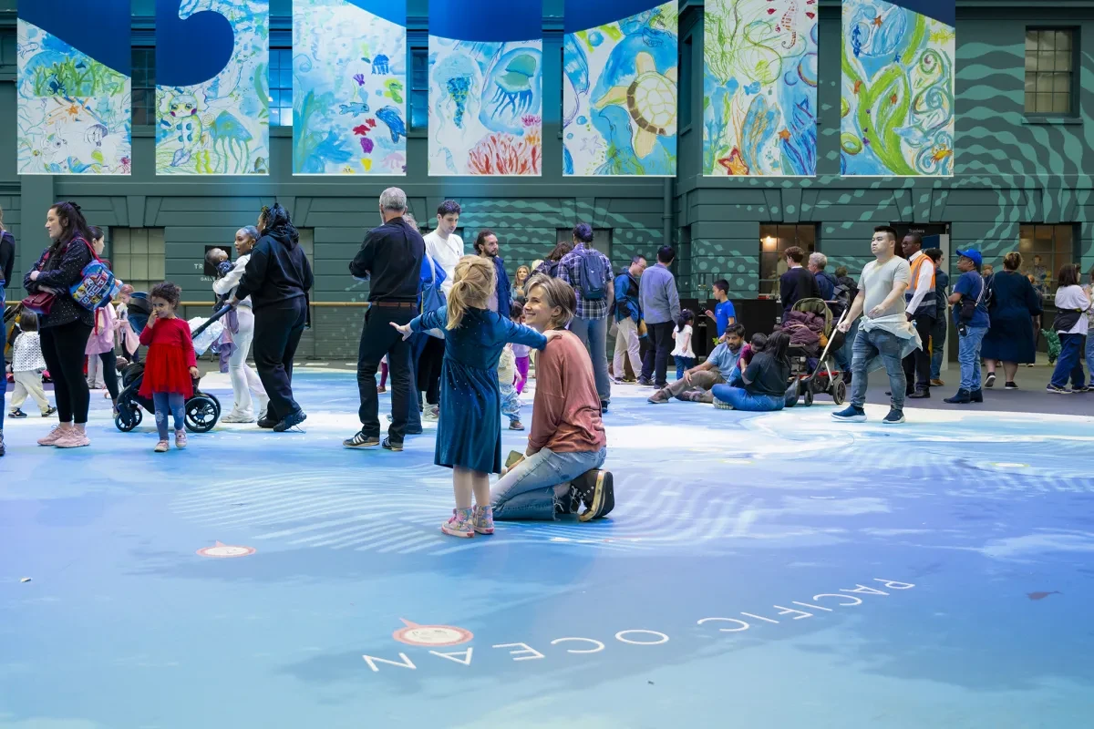 A mother kneels next to her daughter in the centre of the National Maritime Museum's Ocean Map. People pass around them in the background as they enjoy a personal moment in the large space
